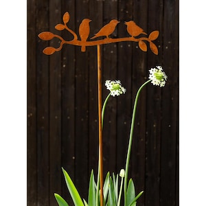Op de afbeelding: Een roestige metalen tuinpaal met een silhouet van een tak en vier vogels. De paal staat in een pot met groene planten en witte bloemen.