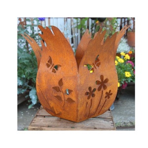 Puede incluir: Una jardinera de metal marrón oxidado con forma de flor, con diseños de mariposas y flores recortadas. La jardinera tiene un aspecto envejecido y está sobre una superficie de madera, con un jardín de fondo.