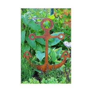 Könnte beinhalten: Ein rostiger Anker aus Metall als Gartenpflock, der aufrecht in einem Gartenbeet mit grünen Pflanzen steht. Der Anker ist eine Silhouette mit abgerundeter Spitze und zwei Armen.