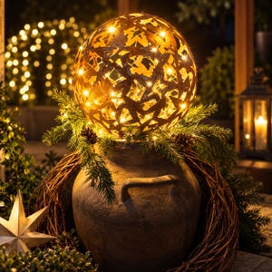 May include: A spherical metal lantern with a star-shaped cutout pattern, illuminated from within by warm white fairy lights. The lantern rests atop a dark, rustic ceramic jar, surrounded by a wreath of pine branches and small pinecones.
