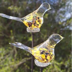 Op de afbeelding: Twee glazen vogelversieringen met geel, bruin en zwart gespikkeld glas. De vogels zitten op metalen palen.