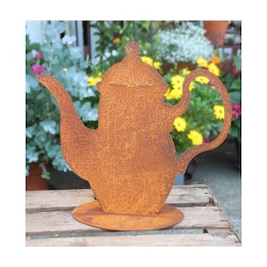 Peut inclure: Une silhouette de théière en métal rouillé avec un corps rond et un bec verseur. La théière est posée sur une base ronde et est placée sur un fond de verdure.