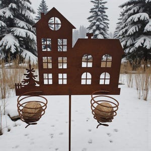K&ouml;nnte beinhalten: Ein rostbrauner Metall-Gartenstecker in Hausform mit ausgeschnittenen Fenstern und einer Baumsilhouette. Zwei spiralf&ouml;rmige Drahtk&ouml;rbe halten runde Steine. Der Stecker steht in einer verschneiten Landschaft mit Tannen im Hintergrund.