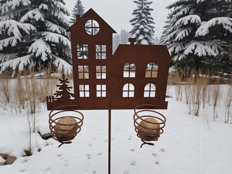 K&ouml;nnte beinhalten: Ein rostbrauner Metall-Gartenstecker in Hausform mit ausgeschnittenen Fenstern und einer Baumsilhouette. Zwei spiralf&ouml;rmige Drahtk&ouml;rbe halten runde Steine. Der Stecker steht in einer verschneiten Landschaft mit Tannen im Hintergrund.
