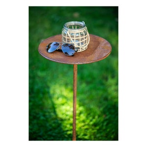 Op de afbeelding: Een roestbruine metalen bijzettafel met een rond blad en een enkel poot. Een witte rieten lantaarn en een zonnebril liggen op het blad.