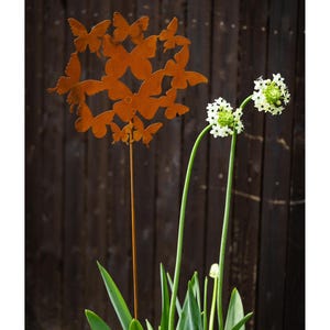 Op de afbeelding: Een metalen tuinpaal met een roestige afwerking heeft een cluster van vlinderuitsnijdingen. De paal is geplaatst in een pot met een groene plant en witte bloemen.