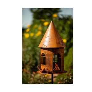 Op de afbeelding: Een roestig metalen vogelhuisje met een puntdak en twee kleine vogels die op de rand zitten. Het vogelhuisje heeft twee ramen en is gemonteerd op een zwarte metalen paal.