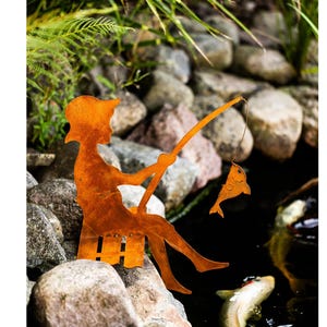 Może przedstawiać: Rdzawa pomarańczowa metalowa rzeźba przedstawiająca osobę łowiącą ryby, siedzącą na ławce. Postać trzyma wędkę z rybą na haczyku. Rzeźba umieszczona jest wśród skał i wody.