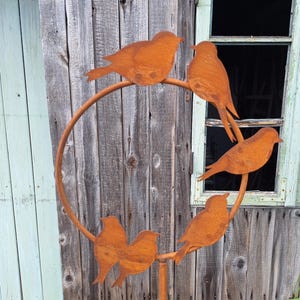 Op de afbeelding: Een roestoranje metalen vogelsculptuur met zes vogels die op een cirkelvormig frame zitten. De vogels zijn gestileerd met eenvoudige vormen en zijn in een decoratief patroon gerangschikt. Op een verweerde houten achtergrond.