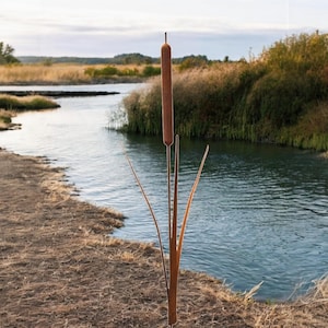 Könnte beinhalten: Eine Metallsculptur einer hohen braunen Rohrkolbenpflanze mit drei langen Blättern. Die Skulptur steht am Rande eines Flussufers mit einem Gewässer im Hintergrund.