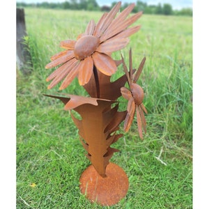 Op de afbeelding: Een roestige metalen bloemensculptuur met twee grote madeliefachtige bloemen en bladeren. De sculptuur, gemaakt van verweerd metaal, staat op een cirkelvormige basis. Het kunstwerk staat buiten in een grasveld.