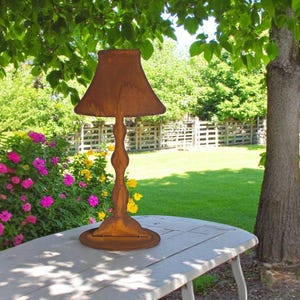 Peut inclure: Une lampe en métal brun rouillé avec un abat-jour, posée sur une table blanche à l'extérieur. La lampe a une tige torsadée décorative et une base ronde. L'arrière-plan présente une pelouse verte, des arbres et une clôture.