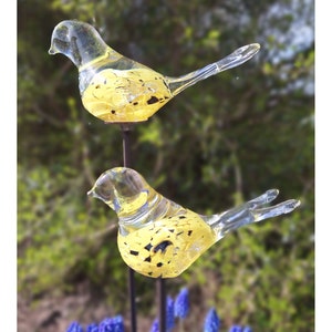 Op de afbeelding: Twee heldere glazen vogels met gele en zwarte gevlekte accenten op metalen stokken. De vogels kijken in verschillende richtingen.