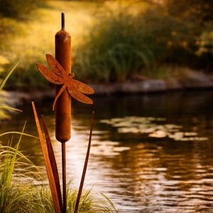 Könnte beinhalten: Eine rostige Metall-Libellen-Skulptur, die an einer Rohrkolbenpflanze befestigt ist, vor einem verschwommenen Hintergrund aus Teich und Blättern. Die Libelle hat detaillierte Flügelmuster. Die Szene erinnert an eine natürliche Umgebung im Freien.