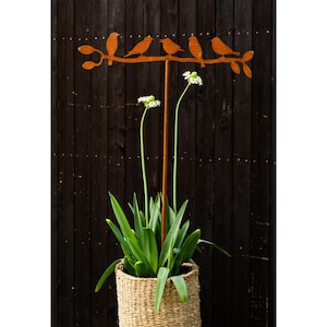 Op de afbeelding: Een roestige metalen tuinpaal met een silhouet van vogels die op een tak zitten. De paal is geplant in een geweven mand met een groene plant.