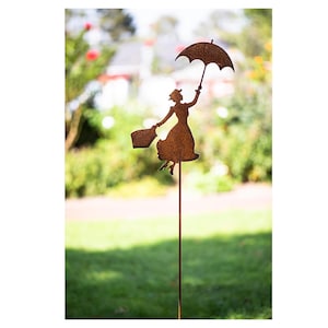 Op de afbeelding: Een roestig metalen silhouet van een vrouw die een paraplu en een mandje vasthoudt, staand op een lange metalen paal. Het silhouet is tegen een wazige achtergrond van groen gras en bomen.