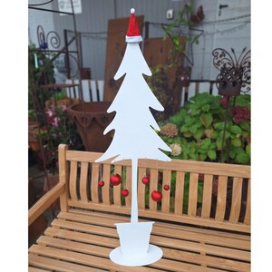 Op de afbeelding: Witte metalen kerstboom met een rode kerstmanmuts op de top en rode kerstballen die aan de takken hangen. De boom staat op een houten bank.