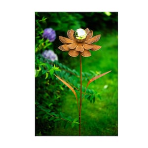 May include: A metal garden stake shaped like a flower with a reflective ball center. The flower petals are a rusty brown color, and the stem and leaves are also brown. The background features green foliage and grass.