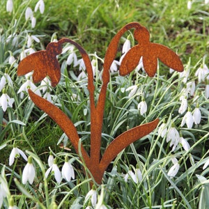 Può includere: Una scultura da giardino in metallo arrugginito con tre fiori di bucaneve stilizzati. La scultura è posizionata in un letto di bucaneve bianchi.