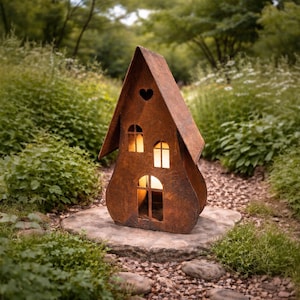 May include: A rust-colored metal lantern shaped like a house with a peaked roof, featuring a small heart-shaped cutout near the top, two arched windows on the upper level, and an arched doorway on the ground floor, all emitting a warm light.