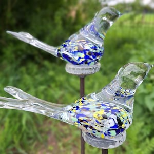 May include: Two glass bird sculptures with a clear body and colourful speckled wings. The birds are perched on metal stakes.