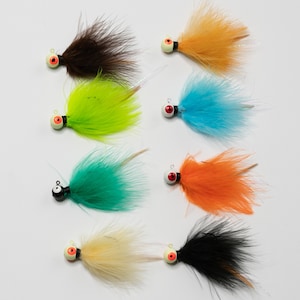 May include: Assortment of eight fishing lures with different colored feathers and black and red eyes. The lures are on a white background.