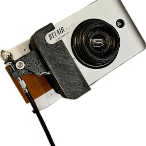Könnte beinhalten: Eine silberne Belair X 6-12 Lomography Kamera mit schwarzem Objektiv und schwarzem Griff. Die Kamera hat einen schwarzen Riemen und eine braun-weiße Basis. Die Kamera ist nach rechts abgewinkelt.