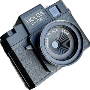 Op de afbeelding: Een zwarte Holga digitale camera met een getextureerde behuizing en een opvallende ronde lensbehuizing. De voorkant van de camera toont de tekst HOLGA DIGITAL boven de lens, terwijl de lensring duidelijk is gemarkeerd met HOLGA OPTICAL LENS.