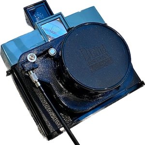 Può includere: Una macchina fotografica istantanea Diana Instant di colore nero e blu con un tappo dell'obiettivo nero. Il tappo dell'obiettivo ha le parole "Diana Instant" in rilievo.