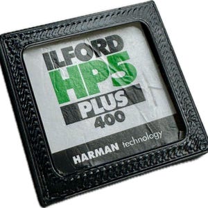 May include: A square black frame with a textured border containing a piece of Ilford HP5 Plus 400 film packaging. The white paper insert features the bold black text ILFORD, large green HP5, white PLUS, black 400, and white HARMAN technology text.