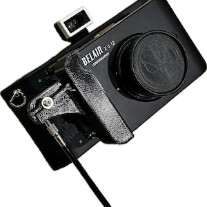 Könnte beinhalten: Eine schwarze Lomography Belair X6-12 Kamera mit Objektivdeckel und Sucher. Die Kamera hat einen schwarzen Gurt und einen silbernen Knopf.