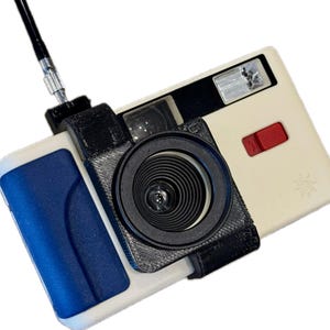 May include: A white and black 35mm film camera with a black lens and a flash. The camera has a blue and white case.
