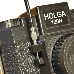 以下が含まれることがあります： 銀色の金属製のノブと黒いレンズが付いた黒いHolga 120Nカメラ。カメラはテクスチャ加工された表面と黒いストラップを持っています。
