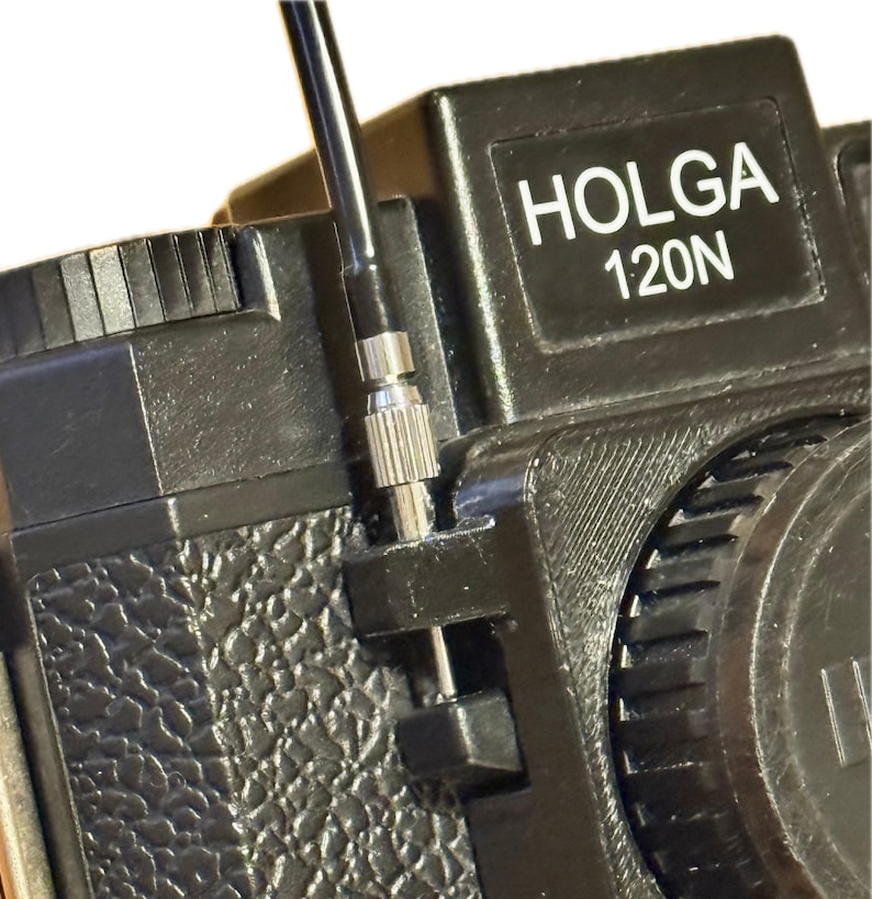 以下が含まれることがあります： 銀色の金属製のノブと黒いレンズが付いた黒いHolga 120Nカメラ。カメラはテクスチャ加工された表面と黒いストラップを持っています。