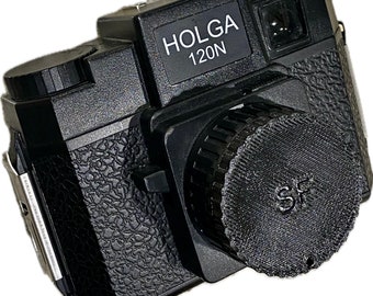 Pode incluir: Câmera de filme Holga 120N preta com corpo texturizado e tampa da lente. A câmera tem formato retangular com flash e lente. A tampa da lente tem as letras "SF".