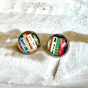 May include: A pair of colorful, round earrings with a silver backing. The earrings feature a collage of colorful patterned paper with text in different languages.