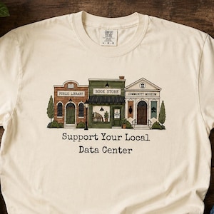 May include: A cream-colored short-sleeve t-shirt featuring a graphic illustration of three buildings labeled Public Library, Book Store, and Community Museum. Below the buildings, the text reads Support Your Local Data Center in black font.