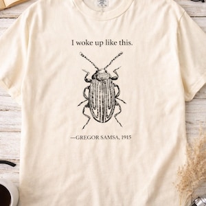 Puede incluir: Camiseta color crema con una ilustración negra de un escarabajo y el texto "I woke up like this." Debajo del escarabajo está el texto "-GREGOR SAMSA, 1915."