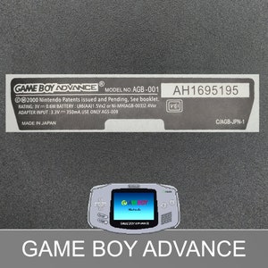 Könnte beinhalten: Eine silberne Game Boy Advance Handheld-Spielkonsole mit einem schwarz-weißen Etikett, auf dem "GAME BOY ADVANCE MODEL NO. AGB-001" und "AH1695195" steht. Das Etikett enthält auch Informationen über die Batterie und den Adaptereingang.