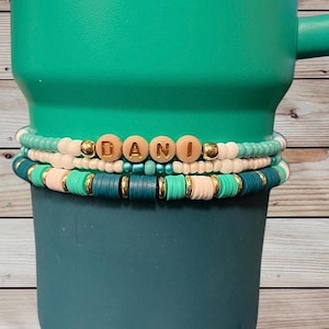 Może przedstawiać: Stos trzech bransoletek z koralików w kolorach turkusowym, różowym, białym i złotym. Górna bransoletka ma drewniane koraliki z literami, które tworzą napis "DANI". Bransoletki są ułożone na turkusowym kubku.
