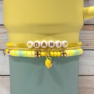 Op de afbeelding: Een stapel gele en witte kralenarmbanden met de naam "DANI" gespeld in witte letterkralen. Een bedel van een gele citroen hangt aan de onderste armband. De armbanden worden weergegeven op een gele en blauwe beker.