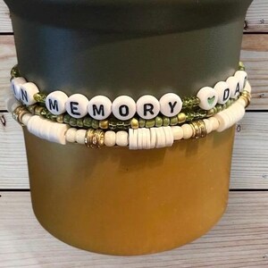 Op de afbeelding: Drie gestapelde armbanden. De bovenste armband heeft witte letterkralen met de tekst "MEMORY DAY" met groene en gouden accenten. De middelste armband heeft groene en gouden kralen. De onderste armband heeft witte en gouden schijfkralen.