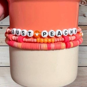 May include: Stack of bracelets in shades of peach, coral, and orange. The top bracelet has white letter beads spelling "JUST PEACHY". The bracelets are stacked on a peach-colored container.