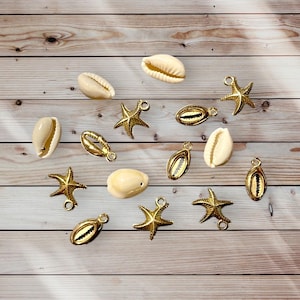 Op de afbeelding: Een assortiment schelp- en zeesterbedels in goud en crème. De bedels zijn verspreid op een lichtbruin houten oppervlak. De zeesterbedels hebben een lus om aan sieraden te bevestigen. De schelpen zijn natuurlijk en hebben een gladde textuur.