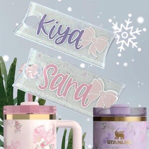May include: Two personalized keychains with the names "Kiya" and "Sara" in cursive script, each paired with a pink bow. The keychains are set against a glittery background. Below are two pink and gold floral Stanley tumblers.