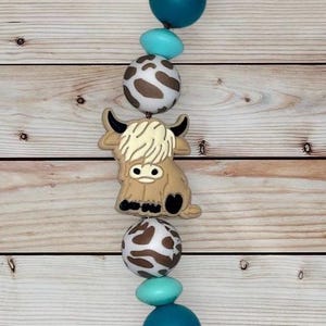 May include: A beaded accessory with a Highland cow charm, teal and light blue beads, and cow print beads. The cow charm is tan with a cream-coloured mane and black horns. The beads are strung on a cord against a wooden background.