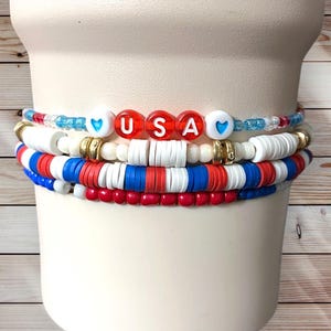 Op de afbeelding: Een stapel patriottische armbanden in rood, wit en blauw. Eén armband spelt "USA" met rode letterkralen. Andere armbanden hebben witte, rode en blauwe schijfkralen en gouden accenten. De armbanden zijn gestapeld op een witte achtergrond.