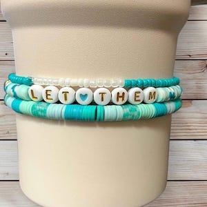 Może przedstawiać: Trzy ułożone na sobie bransoletki z turkusowymi, białymi i niebiesko-zielonymi koralikami. Środkowa bransoletka wymawia "LET THEM" złotymi literami i niebieskim sercem. Bransoletki są prezentowane na białym cylindrycznym kształcie na drewnianym tle.
