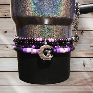 May include: A black tumbler with a glittery, iridescent finish. The tumbler is adorned with several beaded bracelets in shades of purple, black, and pink. A silver charm of a crescent moon with a cat hangs from the bracelets.