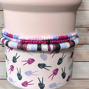 Op de afbeelding: Gestapelde armbanden in roze, blauwe en witte tinten rusten op een lichtroze container. De armbanden hebben een mix van kleine kralen en grotere, platte schijfkralen. De container is versierd met een herhalend patroon van vredestekens.